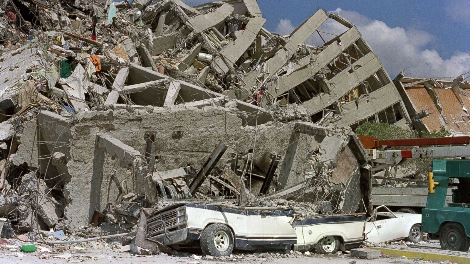 Collapsed building ruins with a crushed white pickup truck in the foreground.