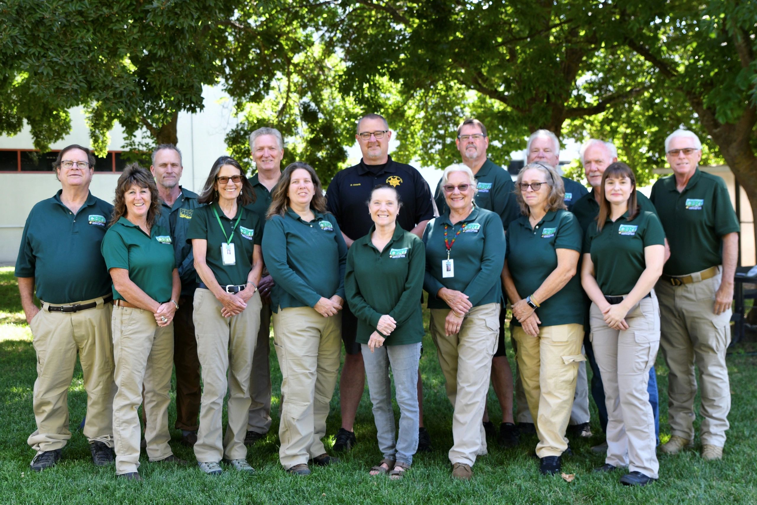 Group photo of 16 people outdoors wearing matching green shirts and beige pants.